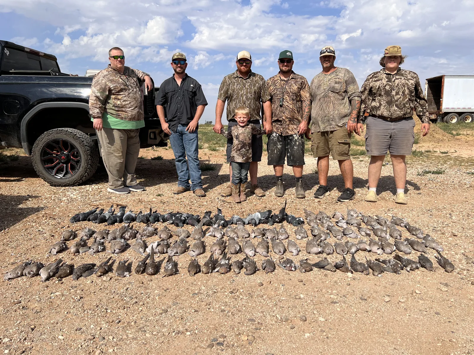 Six hunters and a young boy standing on gravel behind a huge spread of doves laid in rows, a black pickup on one side and a white trailer on the other under a blue sky
