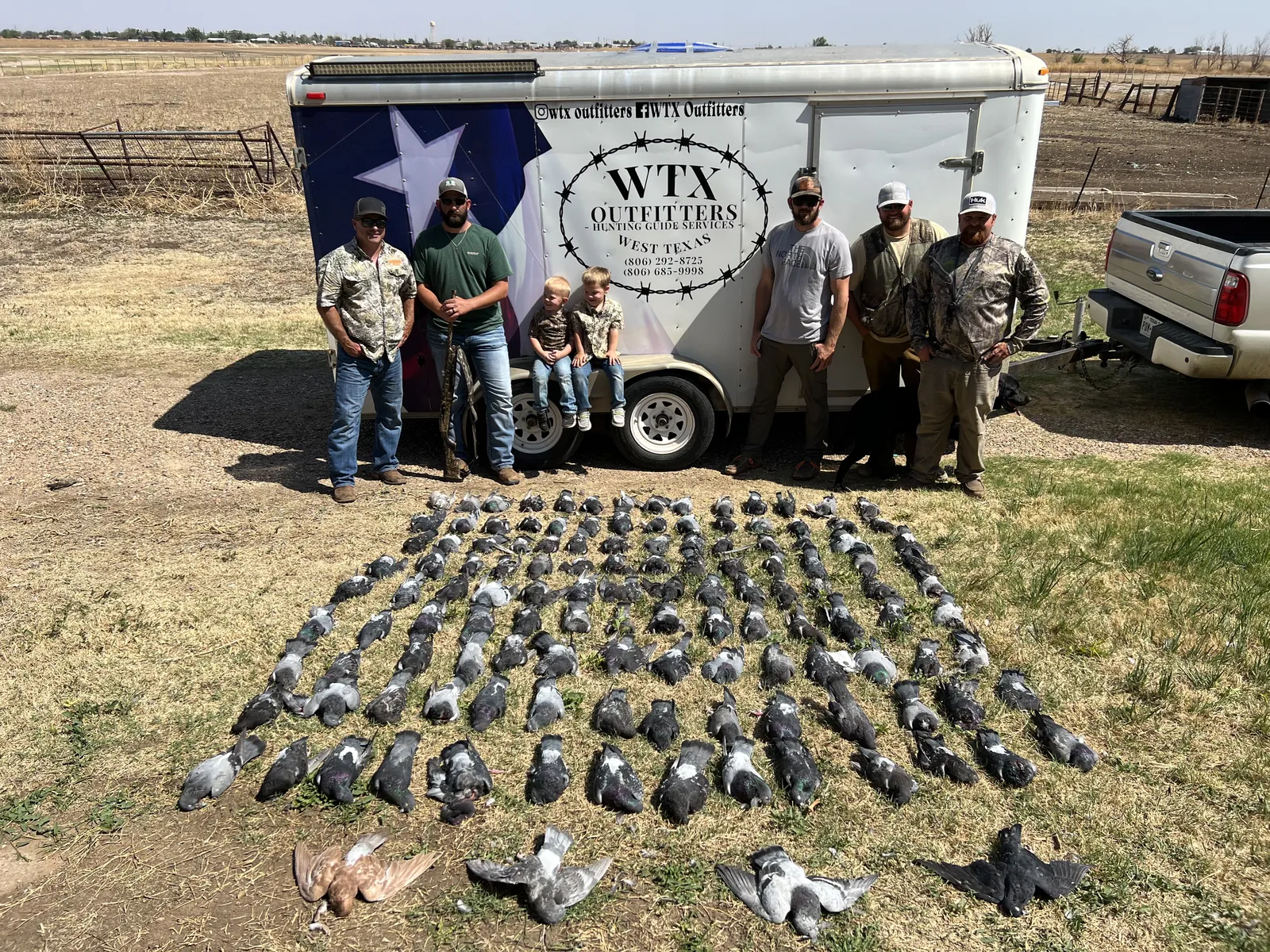 A group of hunters, two young boys and a Labrador at the WTX Outfitters branded trailer behind a huge grid spread of pigeons laid out on the grass